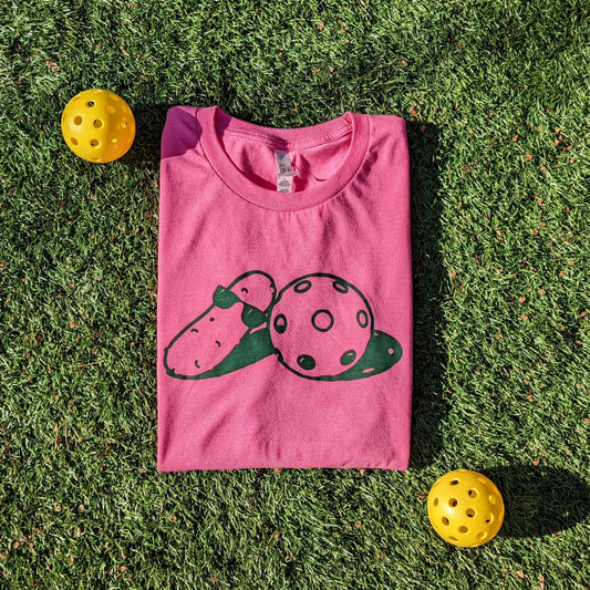 On a bright pink shirt, in emerald green ink, a picture of a pickle drawn with sunglasses next to a drawn pickleball. A pickleball shirt.